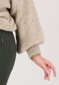 Beige gebreide trui met losse mouwen, geribbelde manchetten en een getextureerde afwerking; olijfgroene broek zichtbaar. Gouden armband met kralen om de pols.