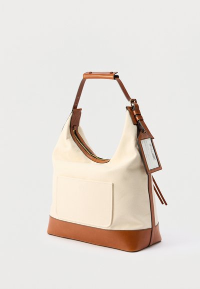 Τσάντα ώμου από καμβά σε κρεμ χρώμα με καφέ δερμάτινη βάση, χερούλι, φερμουάρ και ετικέτα της μάρκας Moschino προσαρτημένη στο πλάι.