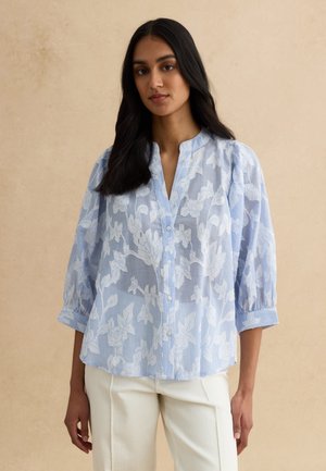 Mujer con cabello largo y oscuro, vestida con una blusa azul y blanca con estampado floral y mangas tres cuartos, y pantalones blancos, sobre un fondo beige.