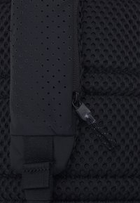 Svart ryggsäck med detaljer som en texturerad meshpanel, dragkedjestängning och en robust rem med en plastdetalj.