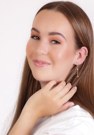 Gouden hangers voorzien van kleine stenen, delicate kettingen en een unieke oorcuff, gedragen door een persoon met steil bruin haar en een lichte huid.