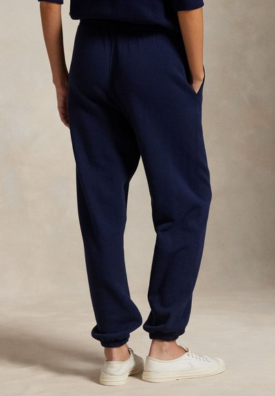 Μπλε ναυτικό παντελόνι φόρμας με ελαστικές μανσέτες, πλαϊνές τσέπες και ελαστική μέση με κορδόνι, συνοδευόμενο από λευκά παπούτσια σε ουδέτερο φόντο.