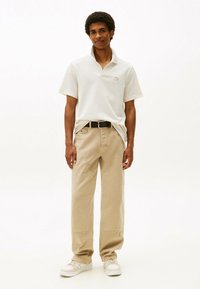 Polo beige con colletto e piccolo logo, abbinato a pantaloni beige larghi. Sneakers bianche completano l'outfit.