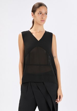 Giovane donna con capelli raccolti che indossa un top nero senza maniche con pannello trasparente e pantaloni neri plissettati, in piedi davanti a uno sfondo bianco.