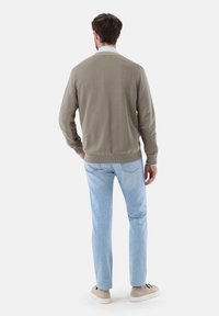 Helle olivfarbene Strickjacke, Rundhalsausschnitt, gerippte Bündchen und Saum, kombiniert mit hellblauen Jeans und beigen Turnschuhen, von hinten betrachtet.