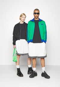 Dwie modelki stoją obok siebie, ubrane w czarne topy, białe szorty z czarnym wykończeniem oraz czarne obuwie; jedna ma na sobie czarną kurtkę, druga zielono-niebieską kurtkę.