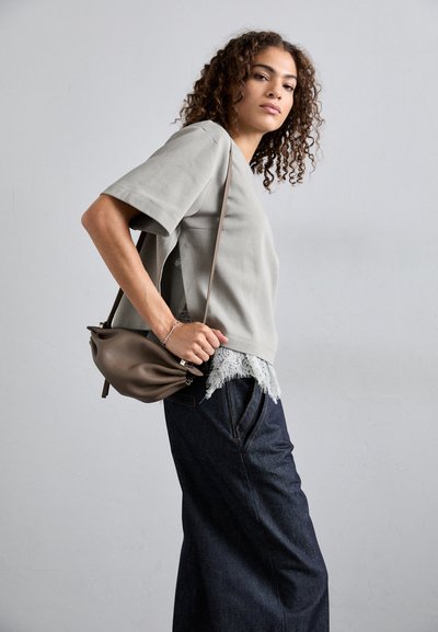 Γκρι κοντομάνικο μπλουζάκι με δαντελένια τελειώματα, συνδυασμένο με σκούρες τζιν παντελόνες σε φαρδύ στιλ. Το μοντέλο κρατά μια μικρή καφέ τσάντα. Απλές, καθαρές γραμμές και ουδέτεροι τόνοι.