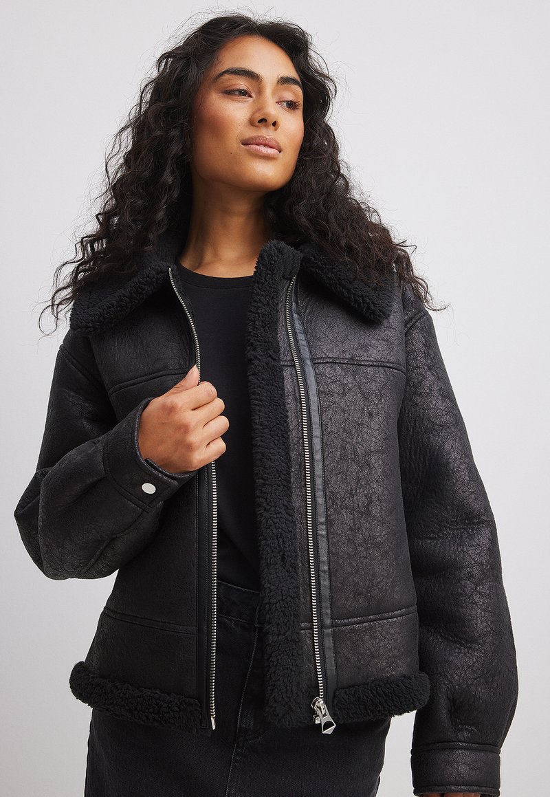 Veste en cuir synthétique noir avec un col et des poignets en fourrure noire. Dotée d'une fermeture éclair et de poches latérales. Surface texturée avec une finition mate.