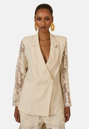 Donna che indossa un blazer doppiopetto crema con maniche trasparenti e testurizzate e pantaloni testurizzati abbinati, accessoriata con grandi orecchini circolari.