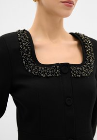 Robe noire avec un décolleté carré orné de perles noires et d'accents dorés, agrémentée d'une fermeture à boutons sur le devant. Texture de tissu lisse.