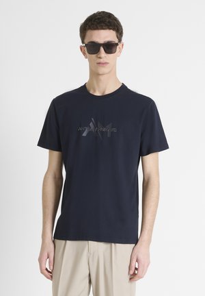 T-shirt en coton bleu marine avec un logo texturé sur le devant. Associée à un pantalon de couleur neutre et des lunettes de soleil noires.