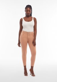 Jean skinny taille haute beige au toucher lisse, avec deux poches avant et une fermeture à bouton. Associé à un débardeur en maille blanche.