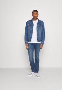 Chaqueta de mezclilla con cierre de botones, dos bolsillos en el pecho, llevada sobre una camiseta blanca, combinada con unos vaqueros azules y zapatillas blancas.