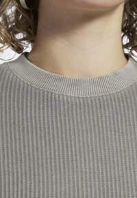 Šedý žebrovaný svetr s kulatým výstřihem. Detailní pohled zdůrazňuje texturovanou tkaninu a pletený vzor, ukazující jemný, ležérní design.