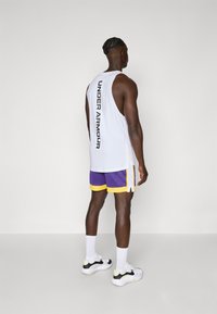 Tricou fără mâneci alb cu "UNDER ARMOUR" imprimat vertical pe spate, asociat cu pantaloni scurți purpurii și adidași alb-negru.