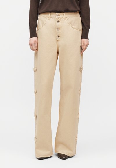 Pantalon beige taille haute à jambes larges avec braguette boutonnée, détails de sangles latérales, porté avec un haut à manches longues marron foncé et des chaussures foncées.