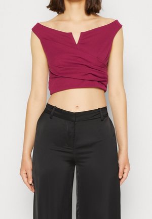 Vrouw met een mouwloze, off-shoulder magenta crop top en hooggetailleerde zwarte wijde broek tegen een effen achtergrond.
