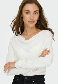 Witte top met lange mouwen en een gedrapeerde halslijn, gemaakt van zachte stof. Het model draagt zwarte jeans en simpele gouden creolen.
