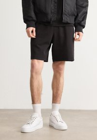 Pantaloni corti neri con una vestibilità rilassata, realizzati in un tessuto morbido e con un orlo che termina appena sopra il ginocchio, abbinati a sneakers bianche e calze a coste.