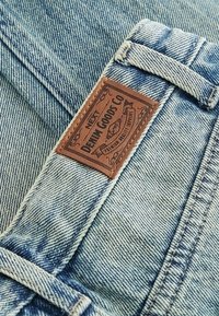 Gros plan d’un jean en denim bleu clair montrant un patch de marque en cuir marron marqué « NEXT Denim Goods Co » près d’une boucle de ceinture.