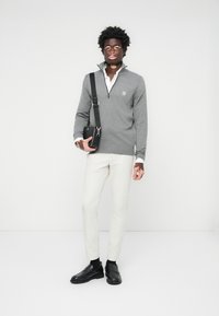 Grauer Reißverschluss-Pullover, darunter ein weißes Hemd mit Kragen, cremefarbene Hose, schwarze Schuhe. Zubehör umfasst eine kleine schwarze Tasche mit einem Riemen.