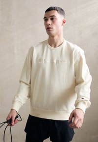 Creme sweatshirt med "Champion" broderet i en matchende farve. Lange ærmer med ribbede manchetter, parret med sorte shorts.