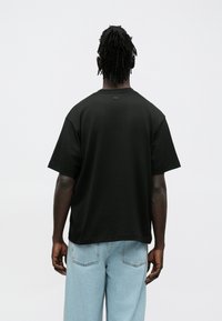 T-shirt preta de manga curta com decote redondo, apresentando uma textura suave e um detalhe discreto de logo nas costas. Combinada com jeans azul claro.