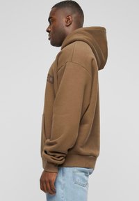 Brauner Hoodie mit einer Kängurutasche und gerippten Bündchen. Hat eine lockere Passform und eine Kapuze mit Kordelzug. Kombiniert mit hellblauen Jeans.