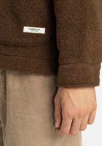 Brązowa polaru z teksturą, z białą metką z napisem "ELEMENT CO. SINCE 1992" na dolnej krawędzi, w połączeniu z jasnobrązowymi spodniami z sztruksu.