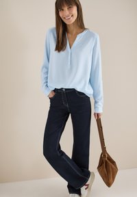 Femme souriante portant un chemisier à rayures bleues claires, un jean foncé, des baskets blanches, tenant un sac à main en daim marron sur un fond beige.