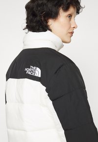 Pufová bunda s čiernou hornou časťou a bielou dolnou časťou, s prešívaním a bielym logom The North Face na ramene.