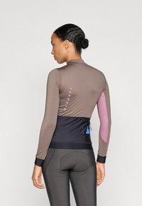 Maglia da ciclismo a maniche lunghe di colore marrone con dettagli in rete rosa. Presenta un design aderente, tessuto testurizzato e logo riflettente sul retro.