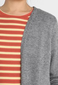 Szary kardigan o miękkiej fakturze zarzucony na czerwono-żółty pasiasty top; ma dekolt w serek i dopasowany krój.