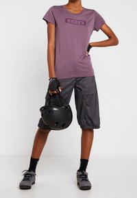 Violetti lyhythihainen t-paita, jossa logo, yhdistetty harmaisiin leveälahkeisiin shortseihin. Vasemmassa kädessä musta kypärä, musta-harmaat jalkineet.