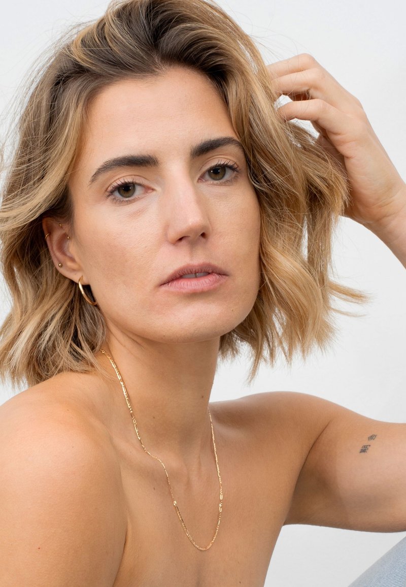 Goldkette mit strukturierten Gliedern, um den Hals getragen; die Schulter einer Person ist sichtbar, mit hellbraunem Haar und minimalem Make-up.