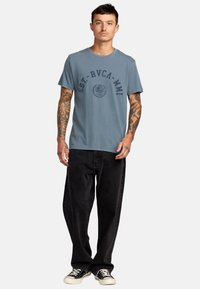 Blaues Baumwoll-T-Shirt mit rundem Halsausschnitt, das ein dunkelbedrucktes Logo und Text auf der Vorderseite zeigt. Kombiniert mit lockeren schwarzen Jeans und Turnschuhen.