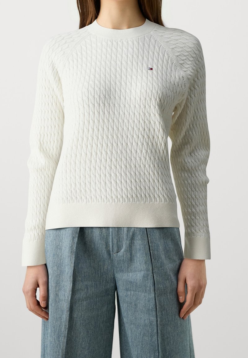 Femme portant un pull en maille torsadée blanche avec un petit logo sur la poitrine et un pantalon plissé ample bleu clair.