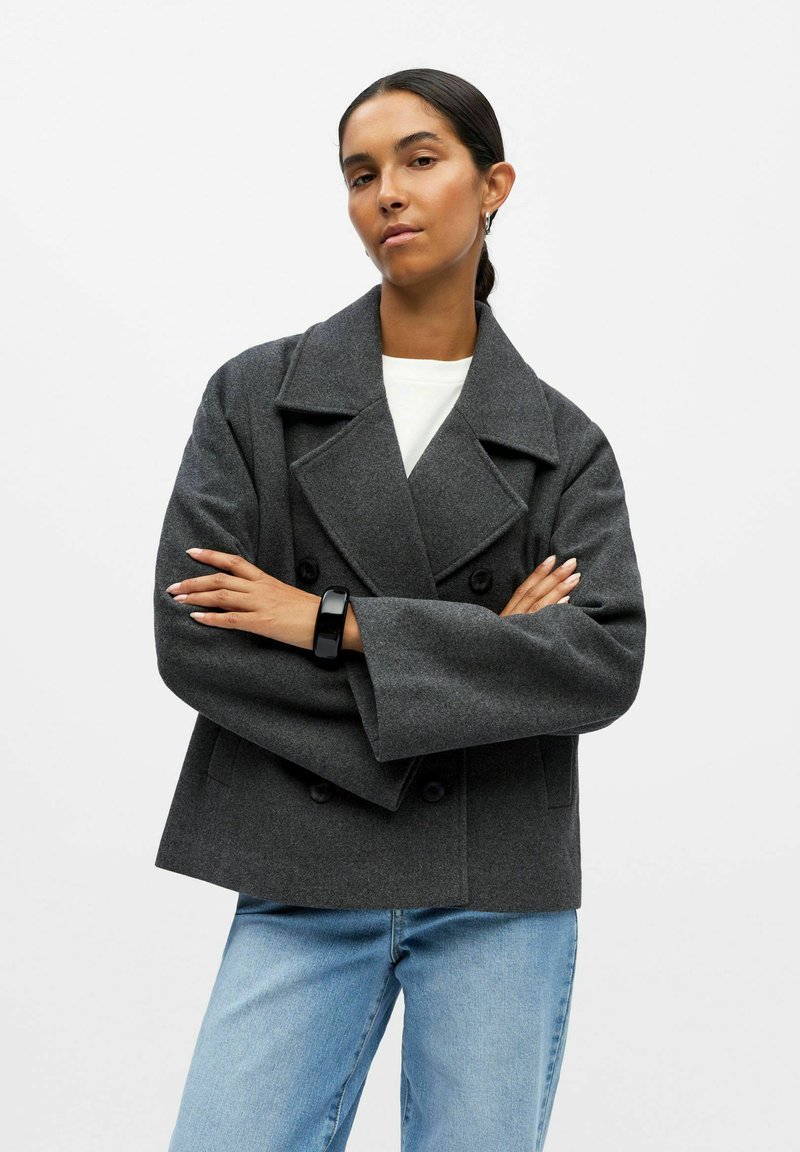 Veste en laine mélangée grise à double boutonnage avec de larges revers, coupe décontractée et grands boutons. Portée sur une chemise blanche et associée à un jean bleu clair.