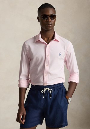 Homme portant des lunettes de soleil, une chemise rose clair à boutons avec manches retroussées, un short bleu marine avec cordon de serrage et une montre-bracelet.