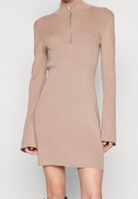 Beigem Rippenstrickkleid mit hohem Kragen und Reißverschlussdetails. Lange Ärmel mit ausgestellten Manschetten, figurnahe Silhouette und mittellange Länge bis zur Oberschenkelmitte.