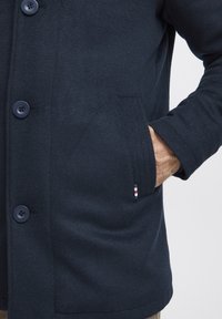 Mornarski vuni kaput s velikim gumbima, bočnim džepom i teksturiranim završetkom. Uključuje mali crveni, bijeli i plavi akcent na džepu.