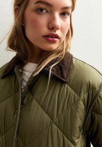 Jong meisje met blond haar, gekleed in een groene gewatteerde jas met een bruin fluwelen kraag, een witte trui en kleine gouden oorringen.