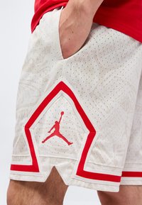 Shorts de basket blancs avec des accents rouges et un logo Jumpman rouge, main reposant dans la poche latérale.