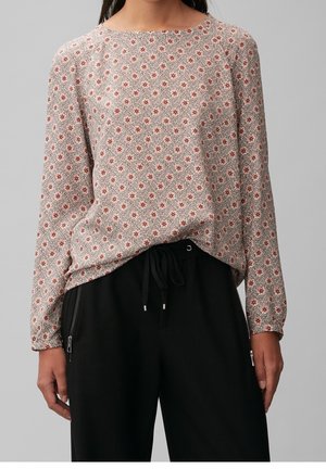 Kobieta ubrana w bluzkę z długim rękawem w mały czerwony kwiatowy wzór oraz czarne spodnie z troczkami i bocznymi kieszeniami na suwak, stojąca na szarym tle.