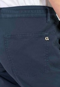 Donkerblauwe broek van een gladde, gestructureerde stof; enkele achterzak met een gouden accentdetail. Strakke lijnen en minimalistisch ontwerp.