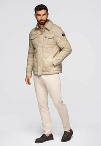 Uomo in piedi con una mano in tasca, indossa una giacca trapuntata beige, pantaloni beige chiaro e scarpe marroni scure su uno sfondo neutro.