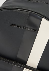 Black nylon backpack featuring a smooth texture, white stripes, and embossed logo on the front. Zipper closure at the top.