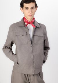 Grå lett jakke med krage, to brystlommer og glidelås foran. Tøyet har en glatt overflate og en skreddersydd passform. Utsmykket med et rødt bandana.