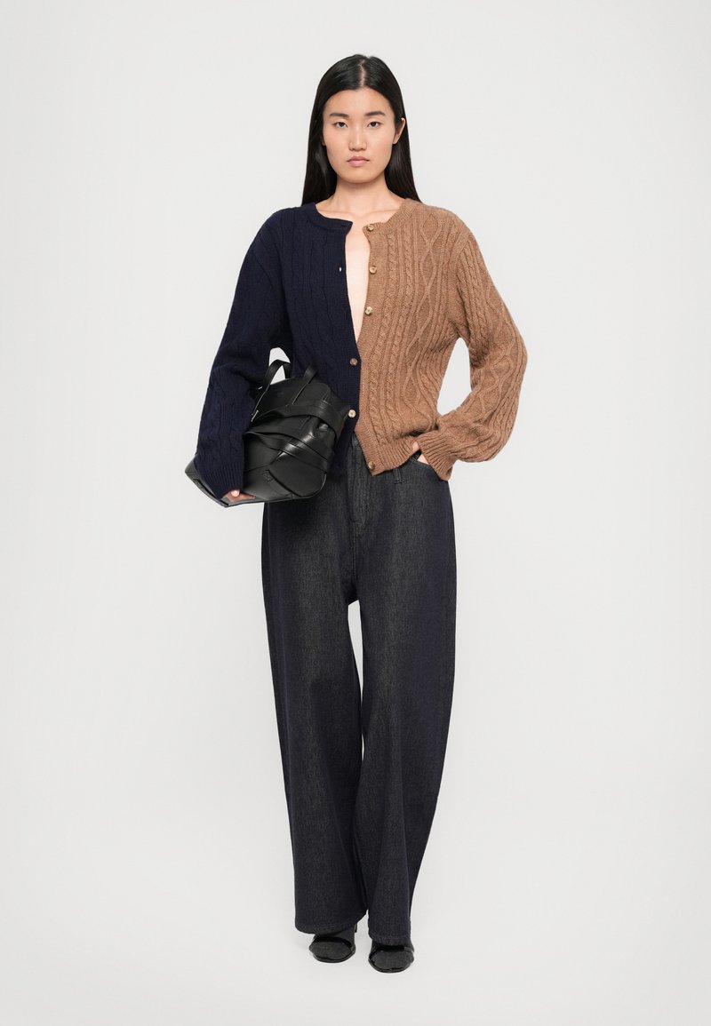 Il cardigan a maglia presenta un design metà marrone e metà blu navy con motivi a treccia. Abbinato a jeans neri a gamba larga e a una borsa nera.