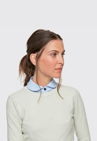 Light green textured sweater with a diamond pattern, layered over a pale blue collared shirt featuring a dark button, worn by a woman.